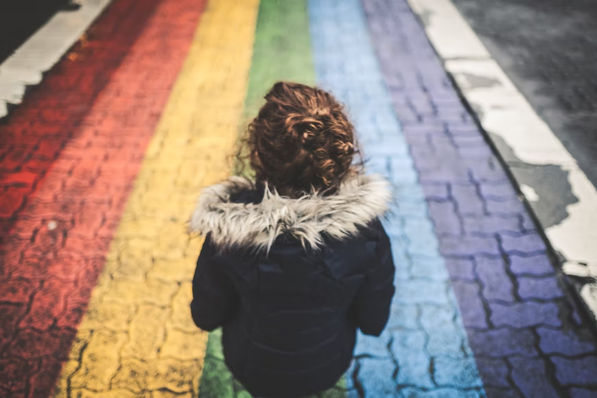 Person standing so you can only see their back. Wearing a black jacket. Standing on a brick road coloured in rainbow stripes.