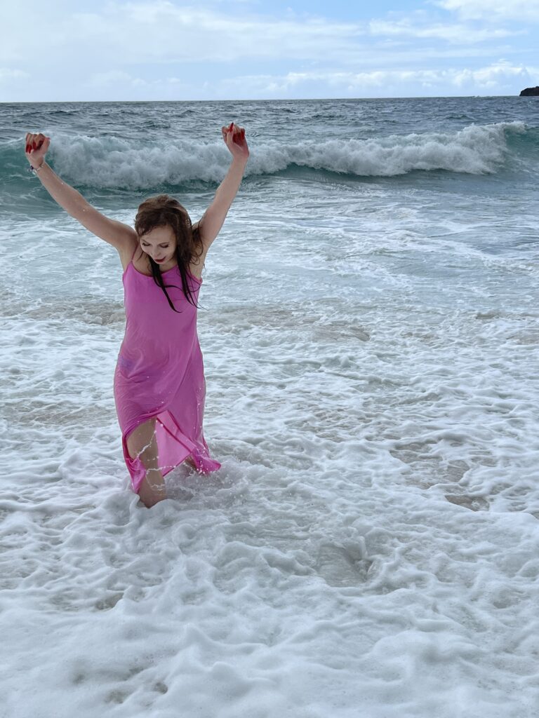 Irene is standing in a pink dress in the water with her arms up in celebration.