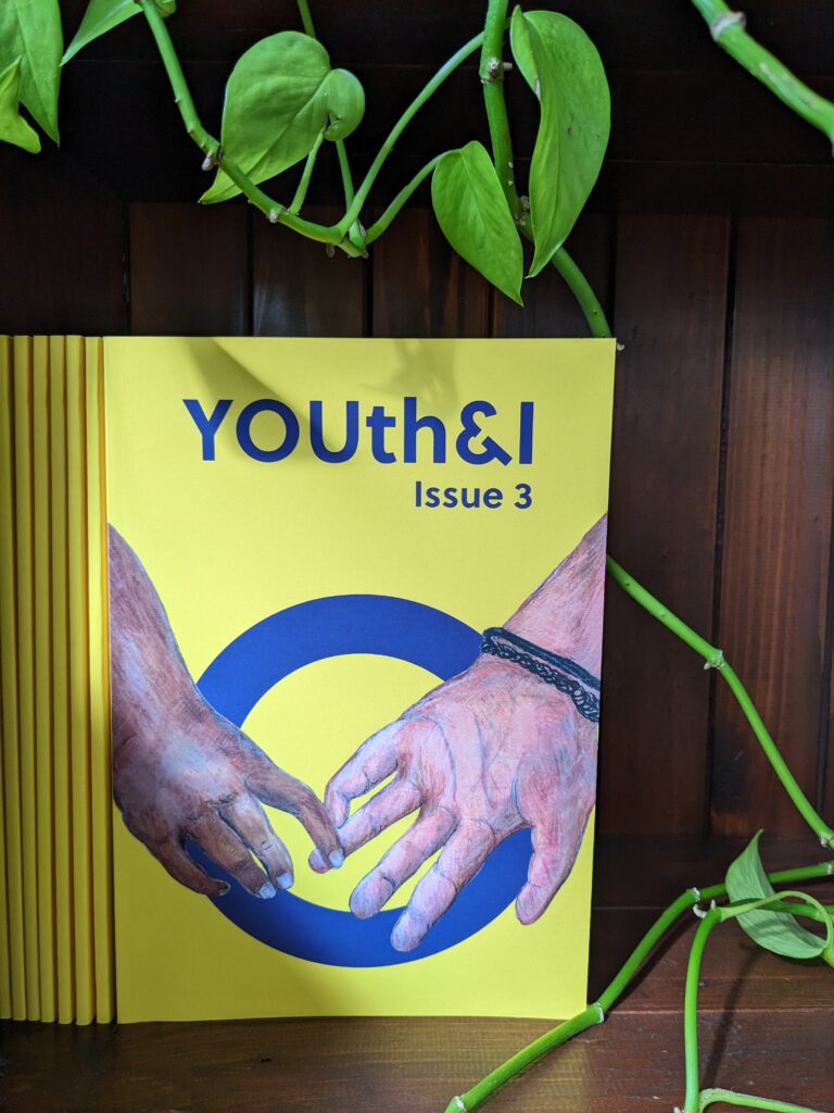 A photo of the front cover of Issue 3 on a brown bookshelf with green vines surrounding the book.