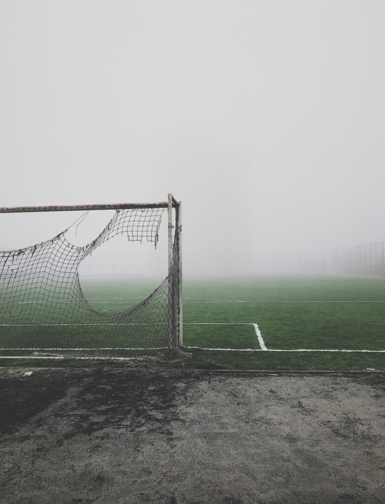 Photo of an empty soccer field in fog. The net in the goal is ripped and falling apart.