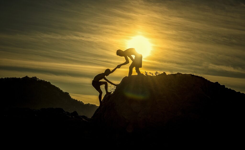 Silhouette image of two people on a mountain top, one helping another climb up. The sun is setting in the background.