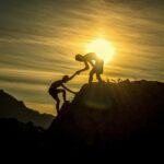 Silhouette image of two people on a mountain top, one helping another climb up. The sun is setting in the background.